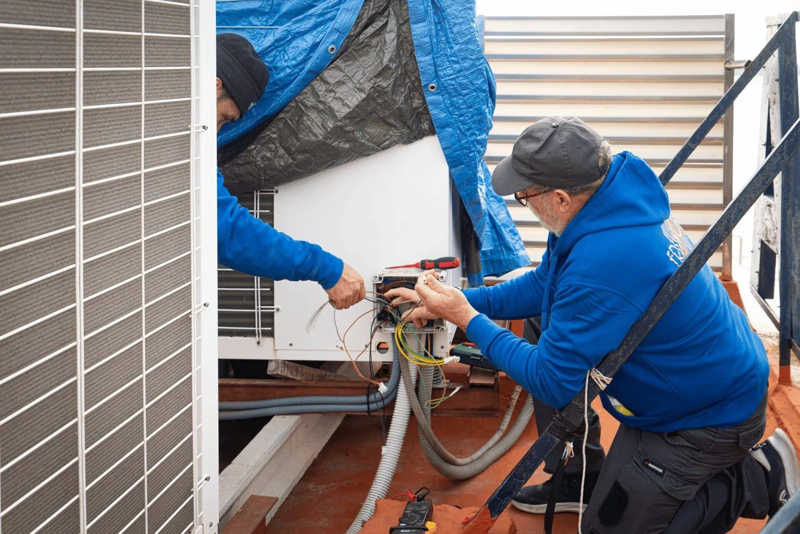 Dos técnicos conectando el cableado de una unidad exterior de climatización en una azotea, imagen propia grabada en las instalaciones de un cliente